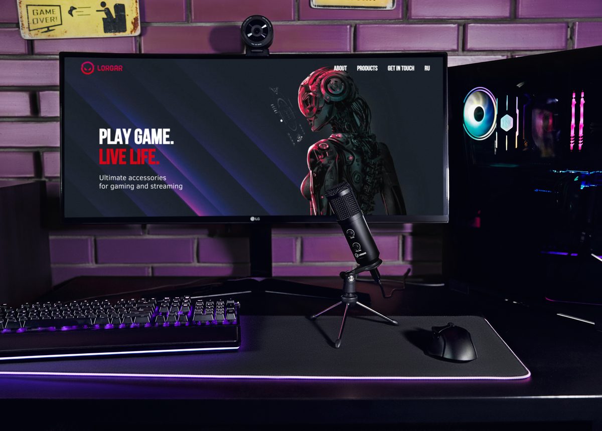 Monitor LORGAR z hasłem Play Game. Live Life. na stanowisku z mikrofonem i podświetlaną klawiaturą