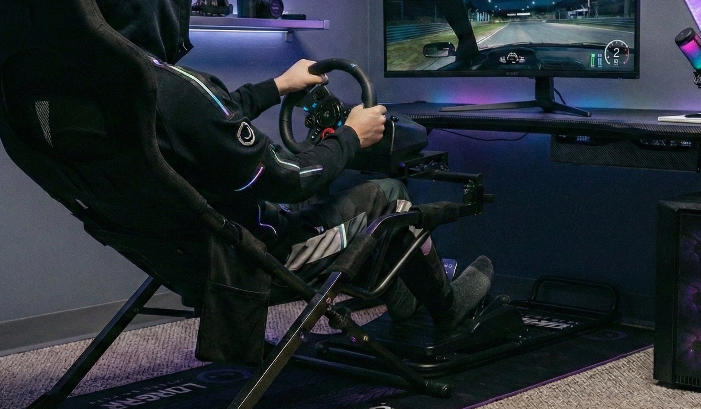 Kierowca trzyma kierownicę kokpitu LORGAR podczas wyścigu na monitorze, na podłodze mata LORGAR Sim Racing